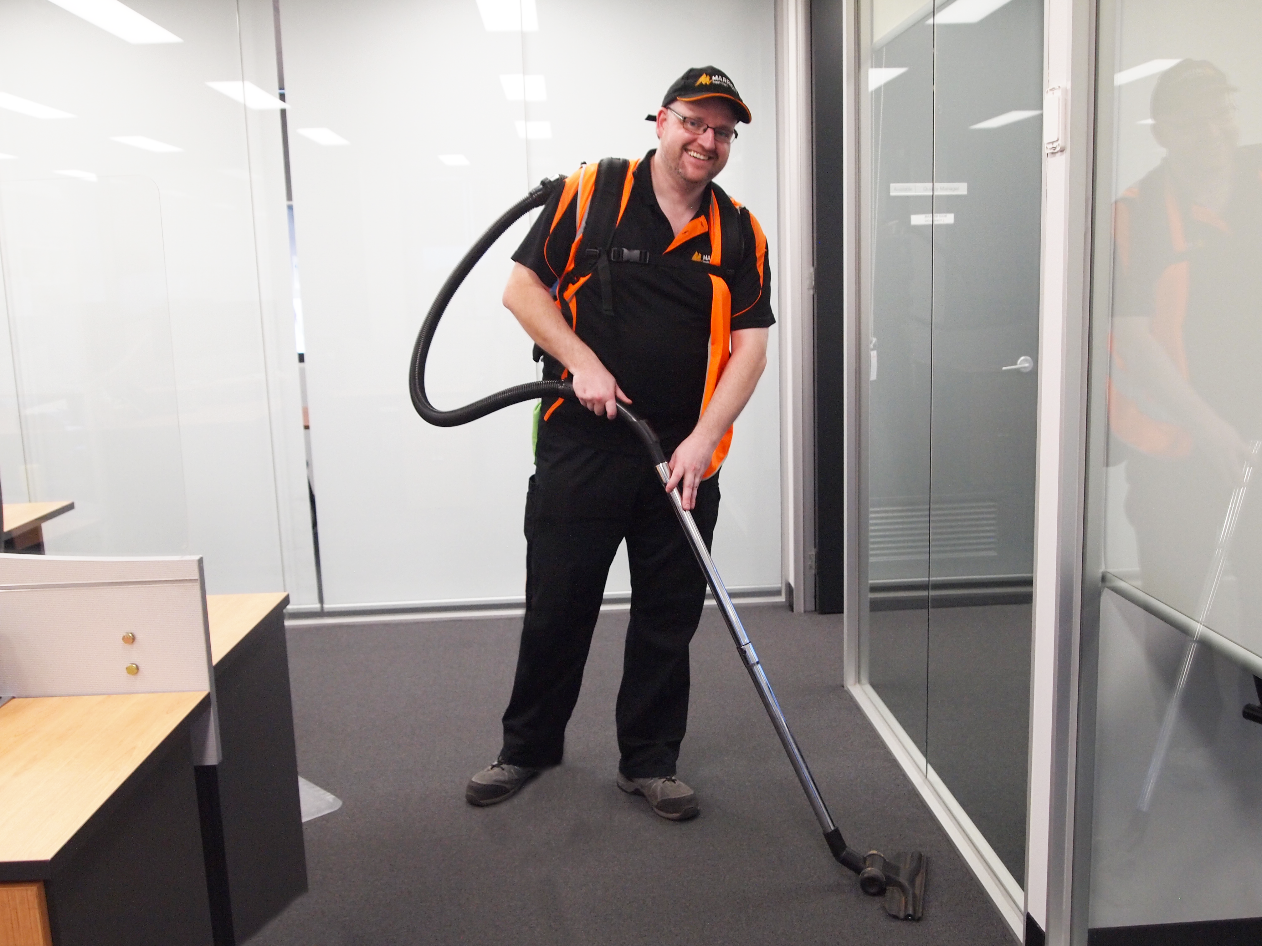 Tall man wearing glasses and smiling whilst vacuuming an office space