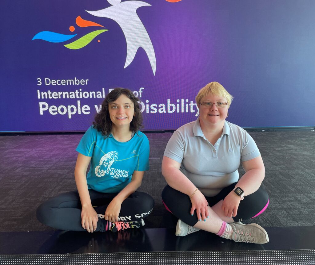 two women with disabilities smiling at the camera and sitting cross legged in front of an international day of people with disability sign